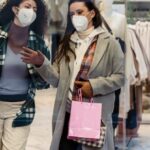 Market Safety - Full body of female friends in protective masks with shopping bags walking through glass doors of clothing store