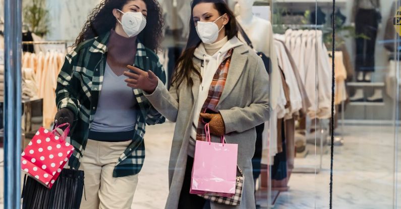 Market Safety - Full body of female friends in protective masks with shopping bags walking through glass doors of clothing store