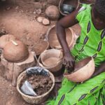 Local Artists - High angle of concentrated African female in light dress sitting on dirty ground of local workshop and creating clay utensils