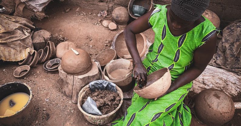 Local Artists - High angle of concentrated African female in light dress sitting on dirty ground of local workshop and creating clay utensils