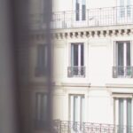 Urban Housing - Hotel with elegant balconies and bay windows in Paris as seen from opposite building through slightly ajar window curtains on sunny day