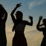 City Events - Low angle silhouettes of unrecognizable young female friends dancing against cloudy sunset sky during open air party