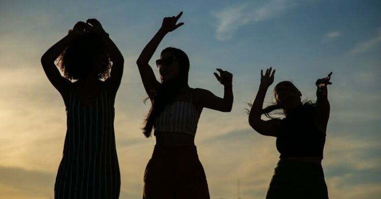 City Events - Low angle silhouettes of unrecognizable young female friends dancing against cloudy sunset sky during open air party