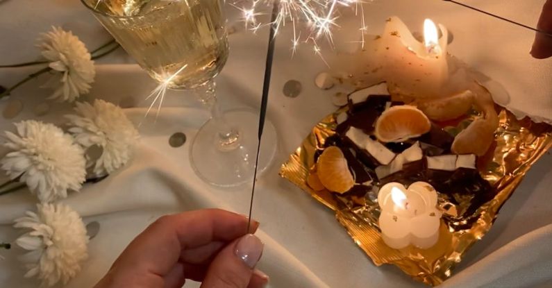 Food Festivals - From above of unrecognizable female with burning Bengal light standing near flowers wineglass and food on tray on white fabric in room with burning candles